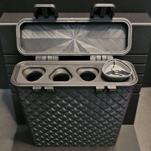 May include: A black rectangular storage case with a textured diamond pattern on the exterior. The lid is open, revealing three empty circular compartments and one grey triangular insert. The interior lid features a geometric starburst texture.