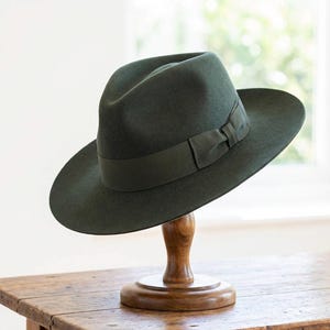 Può includere: Un cappello fedora verde scuro con un nastro e un fiocco in gros-grain abbinati. Il cappello è esposto su un supporto di legno, mostrando il suo stile classico e il design elegante. La tesa del cappello è ampia e la corona è incavata.