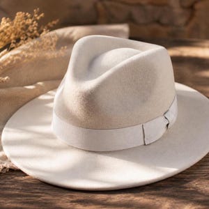 May include: A light beige fedora hat with a wide brim and a matching ribbon band. The hat has a classic teardrop crown and a structured shape. The hat is made of a soft, felt-like material.