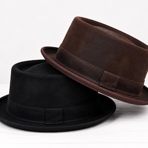 Pode incluir: Dois chapéus fedora, um preto e outro castanho, empilhados. Os chapéus têm um design clássico com a parte superior plana, aba e uma faixa ao redor da coroa. O chapéu castanho está ligeiramente inclinado.