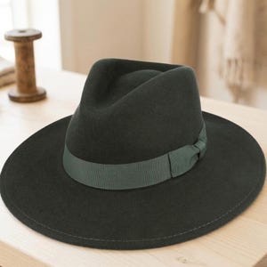 May include: A dark green fedora hat with a wide brim and a matching green ribbon band. The hat is made of a soft material and sits on a light-coloured wooden surface. The hat's design is classic and stylish.