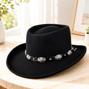 May include: A black cowboy hat with a wide brim and a decorative band with silver-coloured embellishments. The hat is made of a felt-like material and sits on a wooden surface. The hat's design is classic and suitable for various occasions.