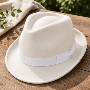 May include: A cream-colored fedora hat with a white band. The hat has a classic teardrop crown and a wide brim. The hat is made of a soft, felt-like material. A stylish accessory for any occasion.