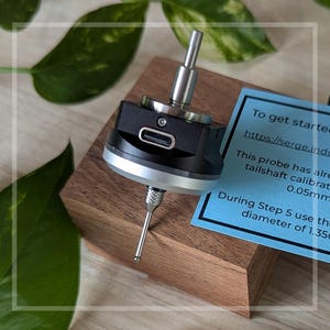 May include: A black and silver probe with a silver tip is mounted on a wooden block. A blue card with white text provides instructions. A clear plastic case and a black cable with a connector are also visible.