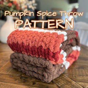 May include: A folded, chunky knit throw blanket in shades of brown, red, white, and orange. The blanket is made of a soft, textured yarn and is labeled "Pumpkin Spice Throw PATTERN". The blanket is folded and stacked on top of another blanket.