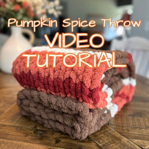 Op de afbeelding: Een gevouwen, dik gebreide deken in tinten bruin, rood, oranje en wit. De deken is op een andere bruine deken gestapeld. Tekst op de afbeelding luidt "Pumpkin Spice Throw VIDEO TUTORIAL".