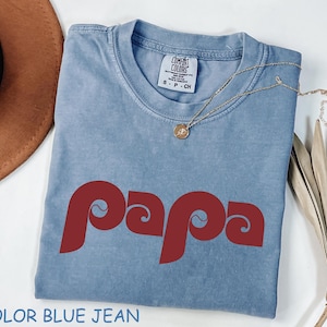 May include: A blue jean colored t-shirt with the word "papa" in a red, retro font. The shirt has a crew neck and short sleeves. A gold necklace with a pendant is draped across the shirt. The text "COLOR BLUE JEAN" is at the bottom.