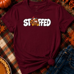 Puede incluir: Una camiseta de manga corta color granate con un gráfico de un pavo marrón con carúncula roja y patas amarillas, situado entre las letras del texto blanco STUFFED, donde el pavo sustituye a la letra U.