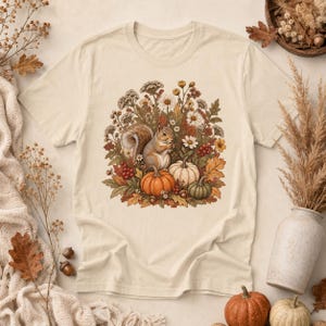 May include: A cream-coloured t-shirt featuring a central graphic illustration of a squirrel surrounded by autumn foliage, including orange and white pumpkins, red berries, brown leaves, and various wildflowers in shades of yellow and white.