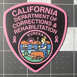 May include: A pink-bordered sticker featuring the words "CALIFORNIA DEPARTMENT OF CORRECTIONS & REHABILITATION" in pink text. The sticker has a black background with a pink ribbon on each side. The center features a state seal with a bear and a woman.