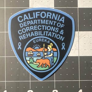 May include: A shield-shaped sticker with the text "CALIFORNIA DEPARTMENT OF CORRECTIONS & REHABILITATION" in light blue. The sticker features a state seal with a bear, a woman, and the word "EUREKA". The border is light blue.