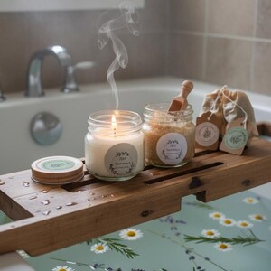 May include: A lit candle with a label that reads "JEM Naturals" sits on a wooden bath tray, next to a jar of bath salts and small linen bags. The candle's smoke rises, and the tub water is decorated with flowers.