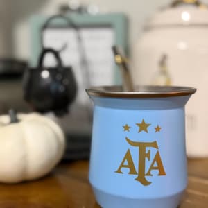 May include: A light blue mate cup with a metallic rim and a gold-colored AFA logo featuring three stars above the letters. A metal straw is inserted into the cup, which has a smooth, curved exterior surface and a wide, open top design.