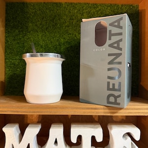 May include: A white mate cup with a silver rim and a metal straw, positioned next to a gray product box labeled REUNATA COLOR. Below the cup and box, large white block letters spell out the word MATE on a wooden shelf surface.