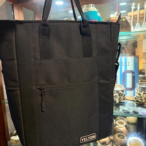May include: A black vertical tote bag featuring two top handles, a front zippered pocket, and a rectangular patch at the bottom right corner with the white text YELTON. The bag has a structured rectangular shape with side panels and black stitching.