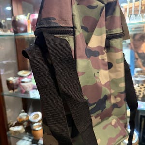 May include: A camouflage print bag featuring a rectangular shape, a black zipper closure on the side, and black nylon webbing straps. The fabric displays a pattern of green, brown, and black organic shapes across the entire exterior surface.