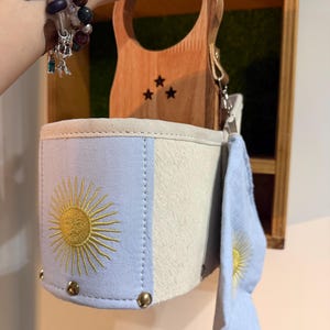 May include: A light blue and cream-colored handbag featuring a yellow embroidered sun design on the front. The bag has a wooden handle with three small star cutouts and a matching light blue fabric accessory attached to the side with a metal clip.