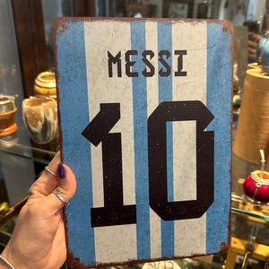 May include: A rectangular metal sign with a distressed finish, featuring vertical blue and white stripes. The sign displays the name "MESSI" above the number "10" in a bold, black font. The sign has a vintage, weathered appearance.