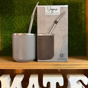 May include: A gray stainless steel mate cup with a matching metal straw, positioned next to its rectangular product box. The box features the text Viajero, ACERO 304, and REUNATA, displaying an illustration of the cup and straw set in a gray tone.