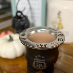 May include: A black mate gourd featuring the AFA logo with three stars on the front and a silver-toned metal rim engraved with various icons, including three world cup trophies, resting on a black stand with a light brown beverage inside.