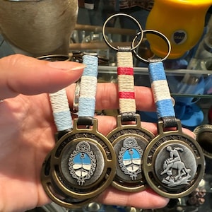 May include: Three Argentinian flag-themed keychains. Each keychain features a circular bronze-colored pendant with the word "ARGENTINA" around the edge and a central emblem. The keychains have blue and white, red and white, and blue and white striped cords.