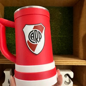 May include: A red beer stein with a white and black logo. The stein has a red handle and a white band around the base. The logo is a shield with the letters "CARP" inside.