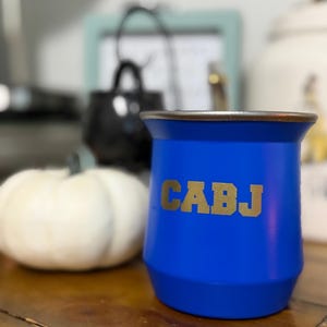 May include: A vibrant blue mate cup featuring a metallic rim and the letters CABJ printed in a bold, gold-toned font across the center. The cup has a wide, flared opening that tapers down toward a narrower base, creating a distinct silhouette.