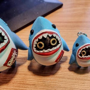 May include: Four shark figurines in varying sizes. The sharks are blue with white bellies, red-lined mouths, and black eyes with gold irises. The largest shark has "Shark" text on its side, and the smallest is a keychain.