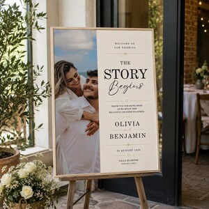 Op de afbeelding: Een rechthoekig welkomstbord voor een bruiloft in een houten lijst met een foto van een koppel aan de linkerkant en crèmekleurige tekst aan de rechterkant. De tekst luidt: WELCOME TO OUR WEDDING, THE STORY BEGINS, OLIVIA and BENJAMIN, 24 AUGUST 2024.