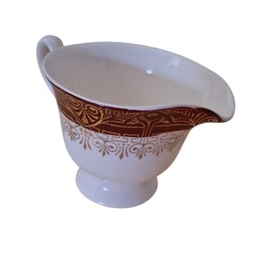 May include: White ceramic gravy boat with a handle and spout. The rim is decorated with a dark red band featuring gold ornate designs. The base of the boat has a pedestal and is also decorated with gold scrollwork.