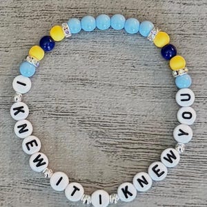 May include: A circular beaded bracelet featuring white letter beads spelling out I KNEW IT I KNEW YOU. The design includes light blue, yellow, and dark blue round beads, interspersed with silver-toned spacers and small clear crystal-encrusted accents.