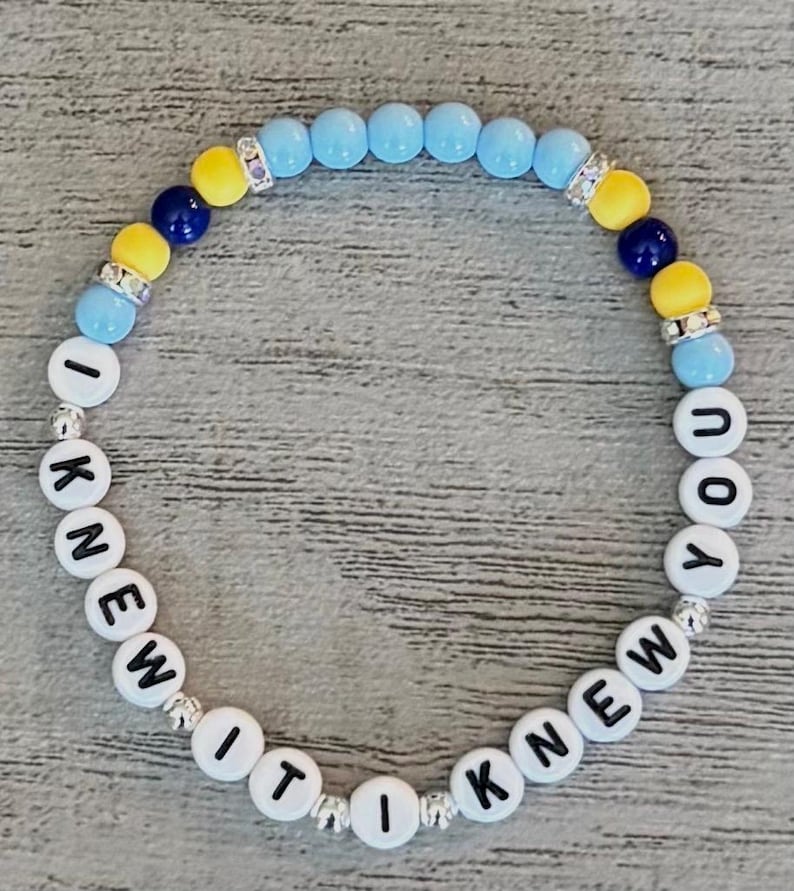 May include: A circular beaded bracelet featuring white letter beads spelling out I KNEW IT I KNEW YOU. The design includes light blue, yellow, and dark blue round beads, interspersed with silver-toned spacers and small clear crystal-encrusted accents.