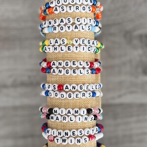 May include: A collection of beaded bracelets featuring team names like "Detroit Tigers," "Houston Astros," and "Los Angeles Dodgers." Each bracelet has white letter beads spelling out the team name, with colorful accent beads. The bracelets are displayed on a tan fabric roll.