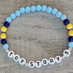 May include: A circular beaded bracelet featuring light blue, dark blue, and yellow round beads. The bracelet includes white letter beads that spell out the text TOY STORY 5, interspersed with small silver-toned metallic spacer beads throughout.