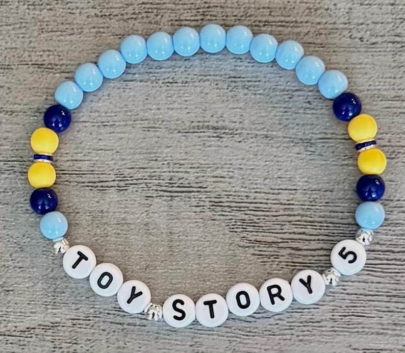 May include: A circular beaded bracelet featuring light blue, dark blue, and yellow round beads. The bracelet includes white letter beads that spell out the text TOY STORY 5, interspersed with small silver-toned metallic spacer beads throughout.