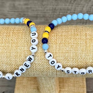 May include: Two beaded bracelets featuring round light blue, dark blue, and yellow beads interspersed with silver-toned spacers. One bracelet displays the white letter beads spelling I KNEW IT I KNEW YOU, while the other spells TOY STORY 5.