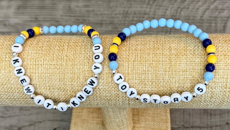 May include: Two beaded bracelets featuring round light blue, dark blue, and yellow beads interspersed with silver-toned spacers. One bracelet displays the white letter beads spelling I KNEW IT I KNEW YOU, while the other spells TOY STORY 5.