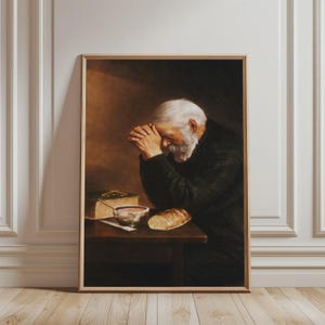 Puede incluir: Impresión enmarcada de un hombre con barba y pelo blancos, con las manos juntas en oración. Una mesa sostiene un libro, un cuenco y pan. La obra de arte tiene una paleta de colores cálidos y apagados.