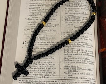 Puede incluir: Rosario negro anudado con cuentas doradas y una cruz, apoyado en un libro abierto con texto del Salmo 83:8. El rosario es un artículo religioso utilizado para la oración y la meditación.