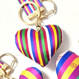 Puede incluir: Un llavero en forma de corazón con rayas verticales en varios colores, incluyendo rojo, verde, amarillo y morado. El llavero tiene un cierre y un anillo dorados. La tela tiene un brillo satinado. El tema general es un accesorio colorido.