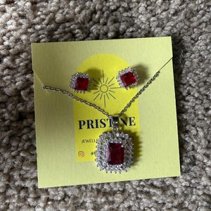 May include: A jewelry set featuring a necklace and matching earrings. The necklace has a square, red gemstone pendant surrounded by clear stones. The earrings also have square, red gemstones. The jewelry is displayed on a yellow card with the text "PRISTINE JEWELLERY".