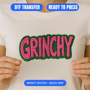May include: A DTF transfer featuring the word "GRINCHY" in pink, edged with green. The transfer is held up against a white backdrop. The words "DTF TRANSFER", "READY TO PRESS", "BRIGHT WHITES", and "QUICK SHIP" are also visible.