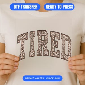 Puede incluir: Una transferencia DTF con la palabra "TIRED" en un diseño de estampado de leopardo marrón y beige. La transferencia la sostiene una persona que lleva una camiseta blanca. La imagen también incluye el texto "READY TO PRESS" y "BRIGHT WHITES • QUICK SHIP".