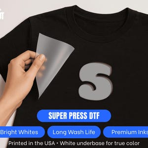 Pu&ograve; includere: T-shirt nero con un design a "S" grigio. Una mano stacca un foglio di trasferimento, rivelando il disegno. L'immagine include il testo: "SUPER PRESS DTF", "Bright Whites", "Long Wash Life", "Premium Inks" e "Printed in the USA".
