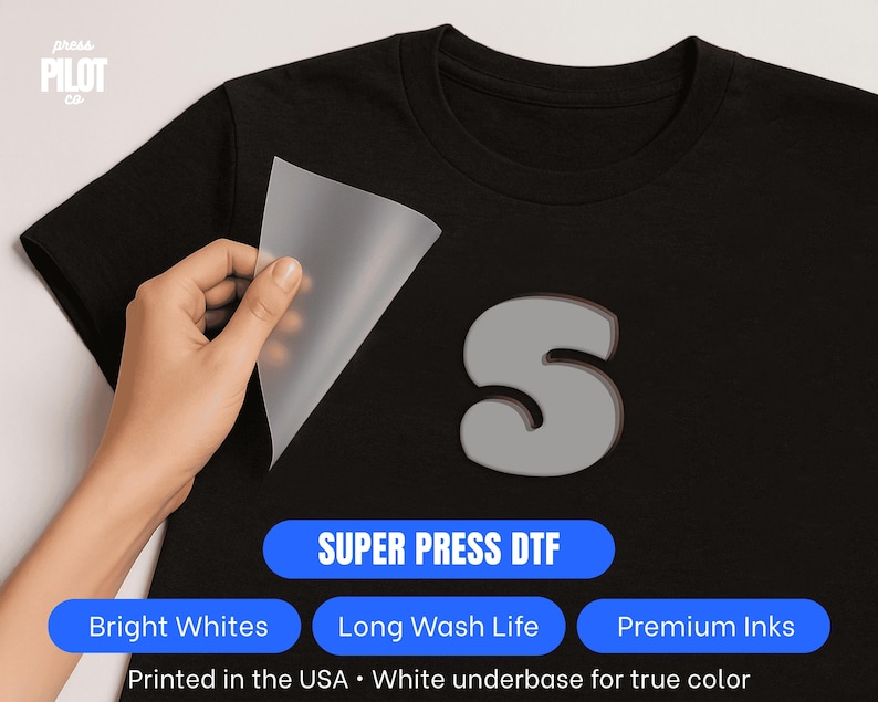 Pu&ograve; includere: T-shirt nero con un design a "S" grigio. Una mano stacca un foglio di trasferimento, rivelando il disegno. L'immagine include il testo: "SUPER PRESS DTF", "Bright Whites", "Long Wash Life", "Premium Inks" e "Printed in the USA".