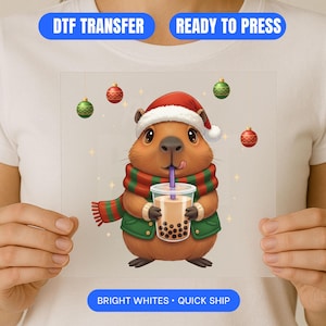 Pode incluir: Uma transferência DTF com um capivara de desenho animado usando um gorro de Papai Noel, cachecol e jaqueta verde, segurando uma bebida boba. O design inclui enfeites de Natal e o texto "DTF TRANSFER", "READY TO PRESS" e "BRIGHT WHITES • QUICK SHIP".