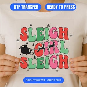 Könnte beinhalten: Ein weißer DTF-Transfer mit dem Text "SLEIGH GIRL SLEIGH" in roten, grünen und pinkfarbenen Buchstaben. Das Design beinhaltet eine schwarze Silhouette von Santa in einem Schlitten, der von Rentieren gezogen wird, mit Schneeflocken und Weihnachtsbäumen. Die Wörter "DTF TRANSFER" und "READY TO PRESS" stehen oben.