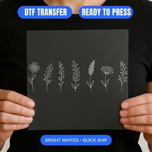 May include: A black DTF transfer sheet with a white floral design. The sheet displays various botanical illustrations, including a daisy, sprigs, and other stylized flowers. The text "DTF TRANSFER" and "READY TO PRESS" are visible at the top, with "BRIGHT WHITES • QUICK SHIP" at the bottom.
