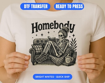 Può includere: Grafica in bianco e nero di uno scheletro che legge un libro mentre è seduto su un divano. La parola "Homebody" è sopra lo scheletro. Una tazza di caffè e una pianta sono accanto al divano. L'immagine è tenuta da una persona che indossa una maglietta bianca.