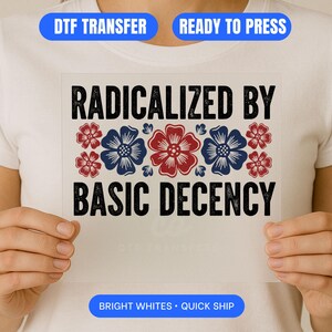 May include: A white DTF transfer with the text "RADICALIZED BY BASIC DECENCY" in black, with red and blue floral accents. The transfer is held up by a person wearing a white t-shirt. The transfer also has the text "DTF TRANSFER", "READY TO PRESS", "BRIGHT WHITES • QUICK SHIP".